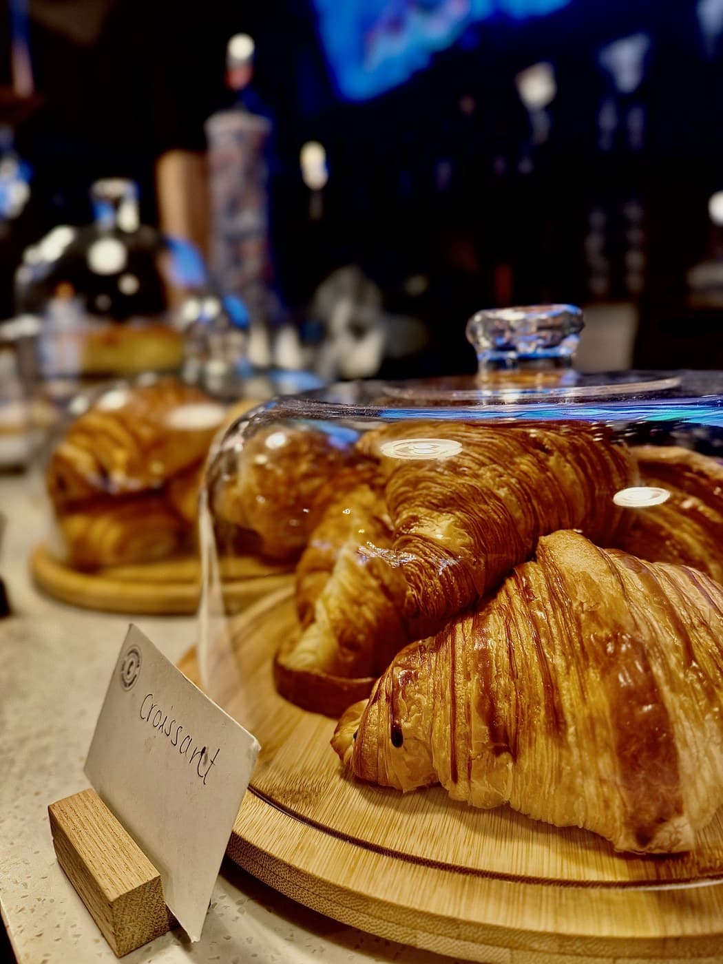 Croissants under a glass cloche on a wooden board at the Oxford Brunch Bar counter, with a handwritten "Croissant" card propped in front